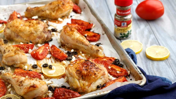 Roasted chicken drumsticks with oven-roasted tomatoes, black olives, lemon slices, and crumbled feta, garnished with oregano and a sprinkle of seasoning.