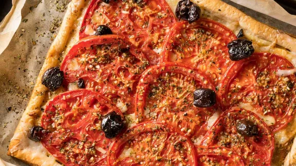 Golden puff pastry tart topped with fresh tomato slices, black olives, and sprinkled with herbs and spices.