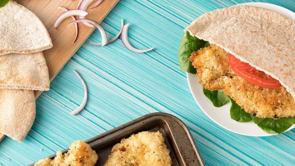 Crispy breaded chicken fillet in whole wheat pita bread with fresh tomato, spinach, and red onion, served with additional pita slices.