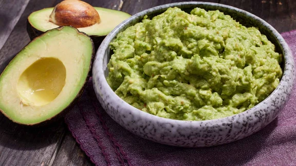Fresh homemade guacamole in a bowl with ripe avocado halves on rustic wooden table.