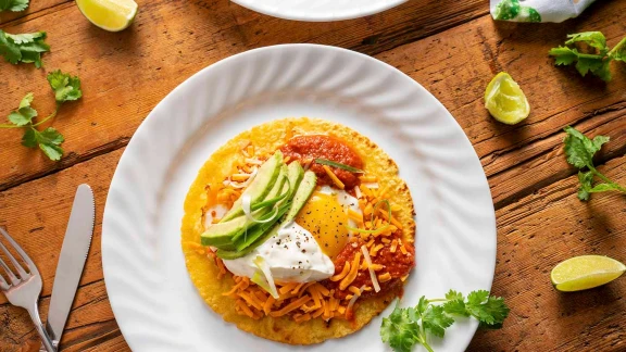 Huevos rancheros with fried egg, shredded cheese, salsa, avocado slices, and sour cream on corn tortilla served on white plate