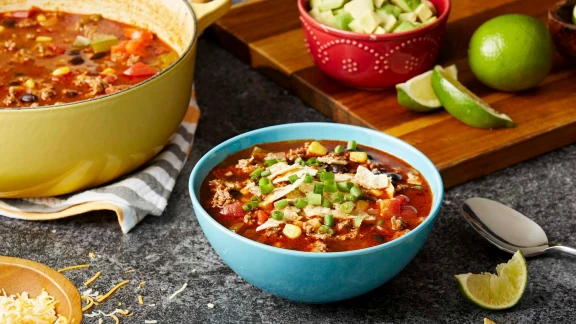 Taco soup with ground beef, corn, tomatoes, black beans, and shredded cheese served in blue bowl with avocado garnish