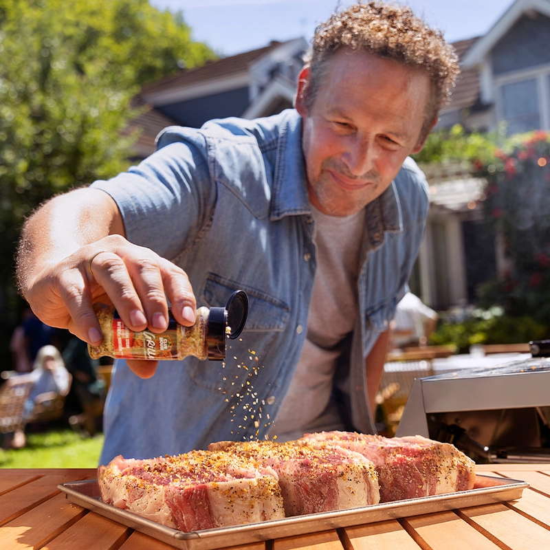 Person sprinkling La Grille seasoning on steaks at a backyard BBQ